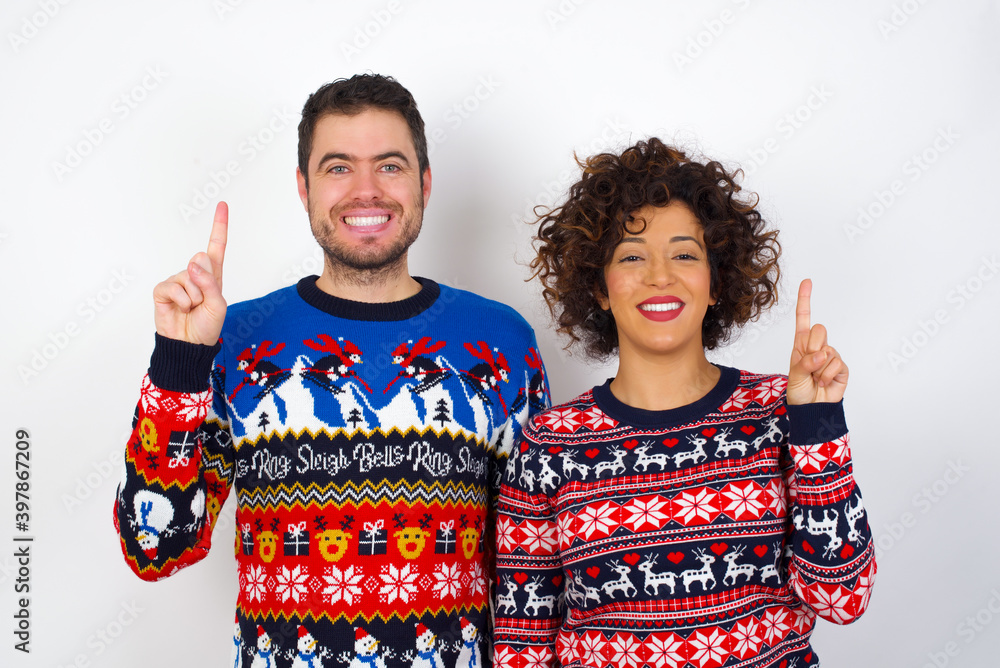 Young couple wearing Christmas sweater standing against white wall showing and pointing up with finger number one while smiling confident and happy.