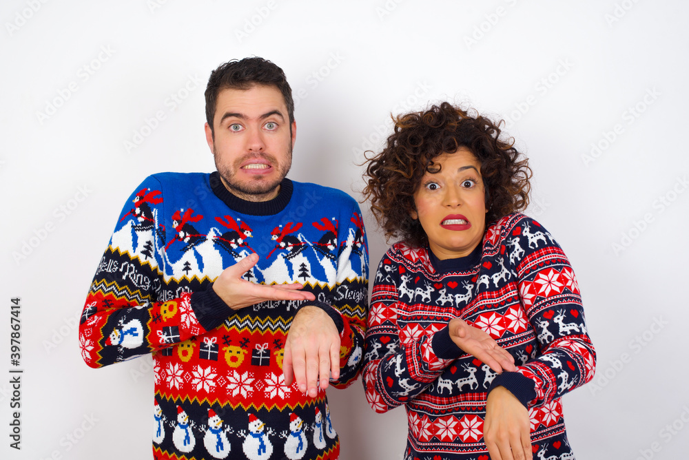 Young couple wearing Christmas sweater standing against white wall In hurry pointing to watch time, impatience, upset and angry for deadline delay.