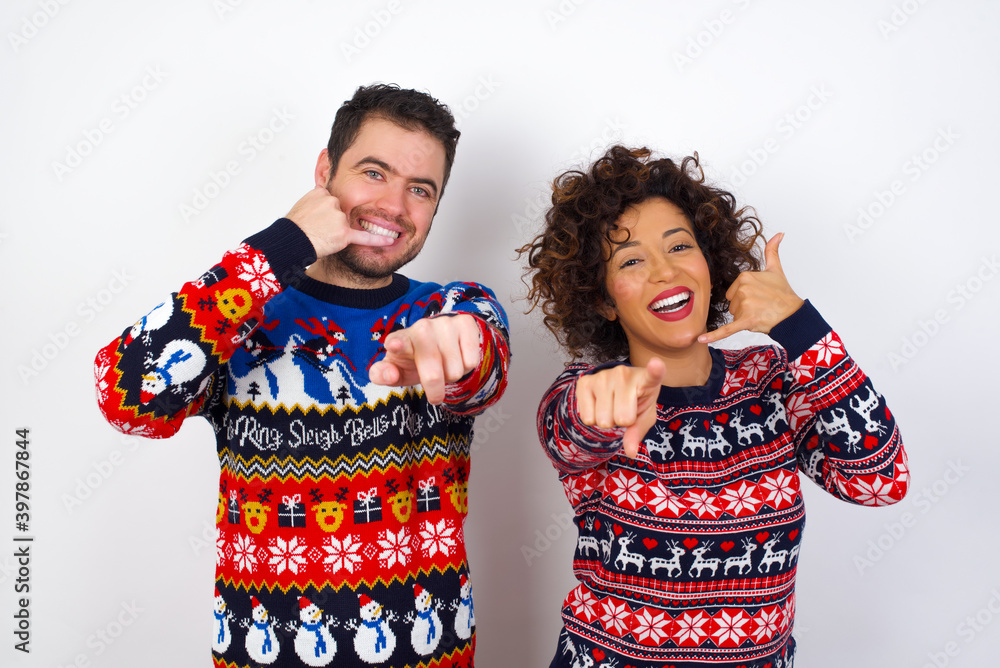 Young couple wearing Christmas sweater standing against white wall smiling cheerfully and pointing to camera while making a call you later gesture, talking on phone