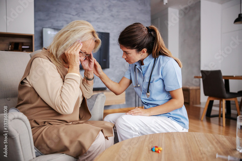 Blond senior woman sitting at home and holding head because she s having headache. Nurse sitting next to her and comforting her.