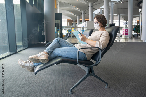 Young female freelancer wearing protective medical mask sits in comfortable chair in airport lounge and works using digital tablet, ready to travel. Life and travel during pandemic