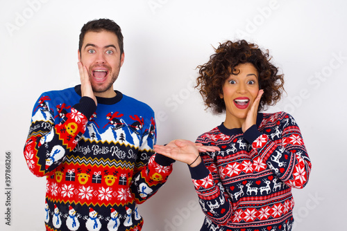 Crazy Young couple wearing Christmas sweater standing against white wall advising discount prices hold open palm new product