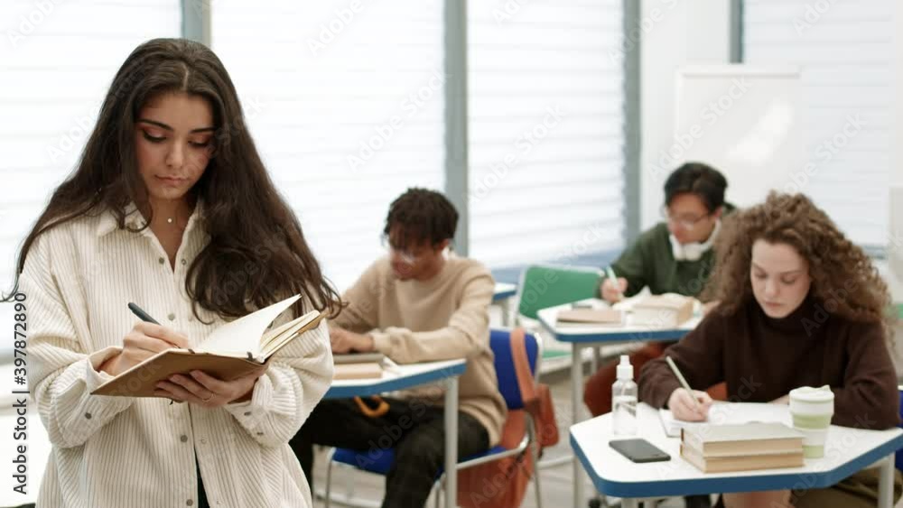 Medium POV of young Mixed-Race woman taking notes in workbook, then closing it, looking up and smiling on camera, standing in front of blurred students sitting by desks, writing in classroom Medium POV of young Mixed-Race woman taking notes in workbook, then closing it, looking up and smiling on camera, standing in front of blurred students sitting by desks, writing in classroom