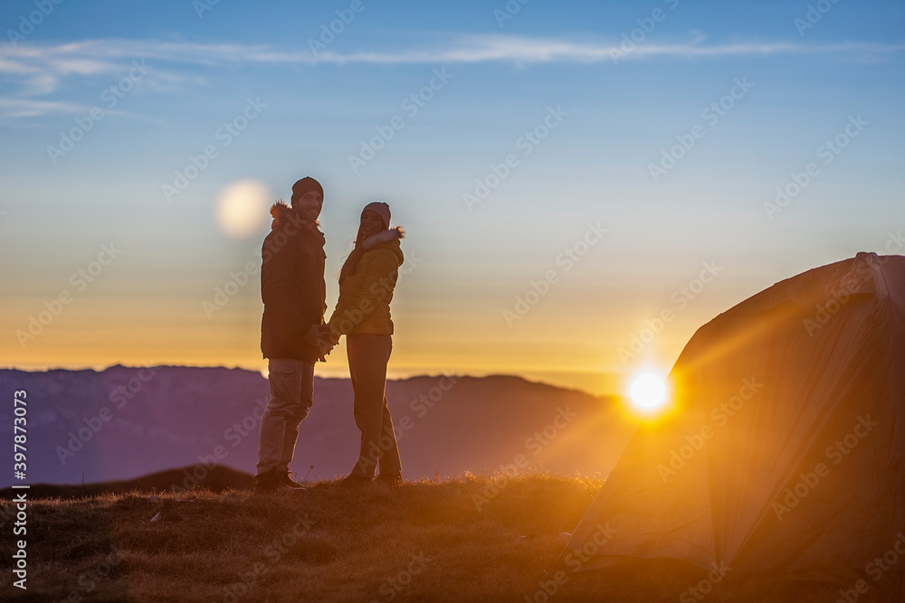 A young couple in front of the tent is holding hands and enjoying the sunset at the top of the mountain. 
