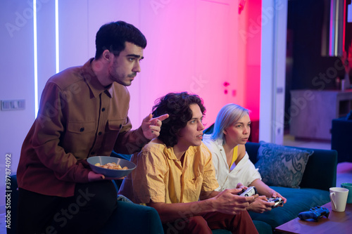 Happy young people playing video games on console while sitting on couch in front of tv. Man advice his friend in a game. Room with warm and neon lights.