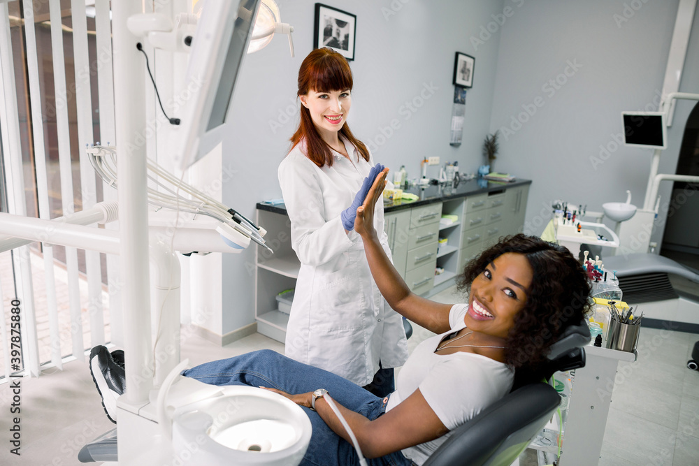 Fototapeta premium Dental caries prevention concept. Satisfied African woman patient at the dentist's chair gives five to her cheerful female dentist after treatment. Focus on Caucasian woman dentist