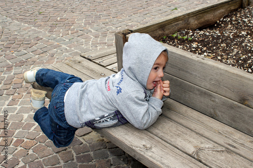 Un niño pequeño con capucha tumbado en un banco de madera jugando con las manos en la boca.