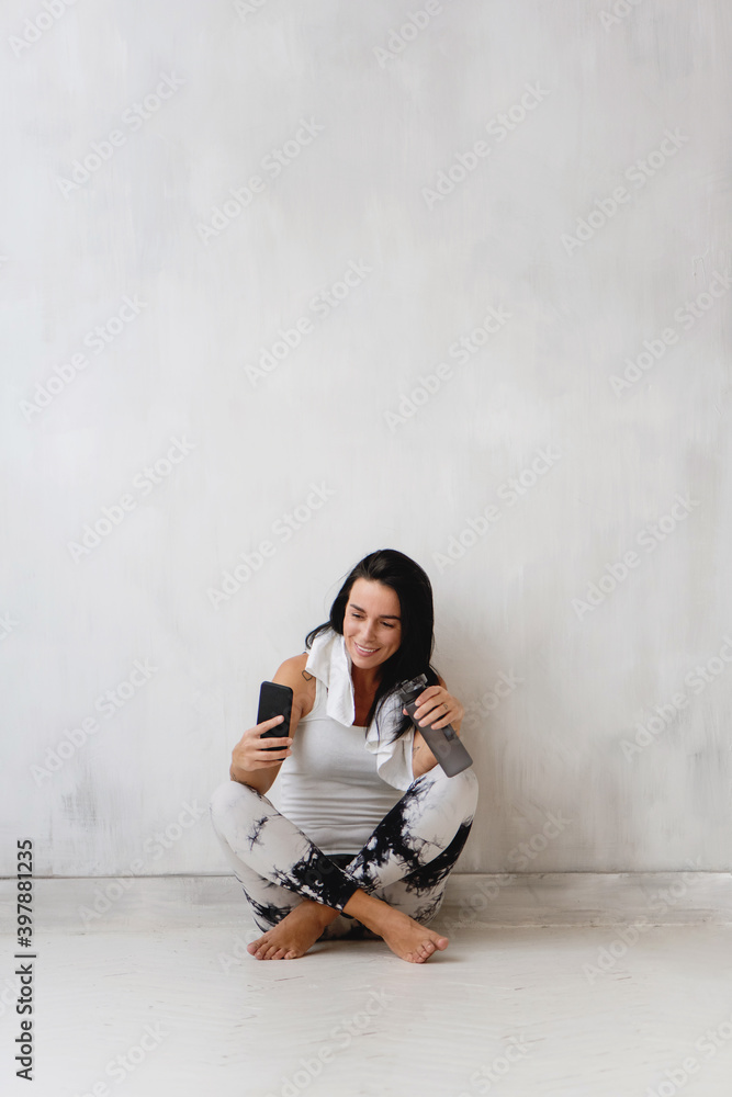 Woman sitting on the floor and doing selfie after workout on the background of white wall.
