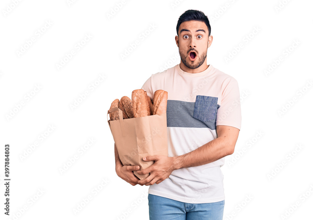 Young handsome man holding bread bag scared and amazed with open mouth for surprise, disbelief face Young handsome man holding bread bag scared and amazed with open mouth for surprise, disbelief face