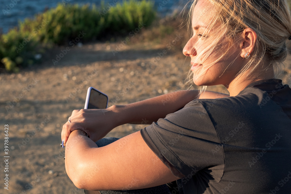 Blond woman with face mask on her back looking at the mobile at sunset
