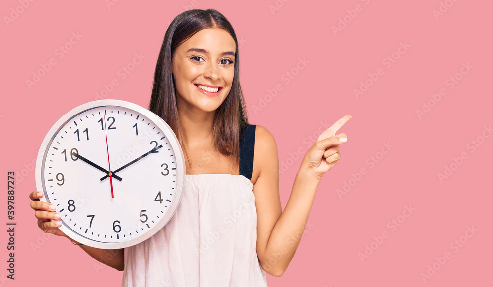 Young hispanic woman holding big clock smiling happy pointing with hand and finger to the side