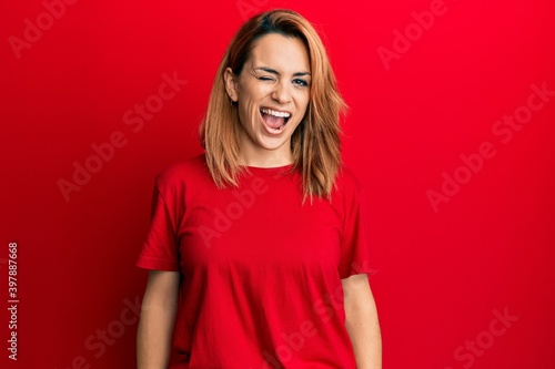 Hispanic young woman wearing casual red t shirt winking looking at the camera with sexy expression, cheerful and happy face.