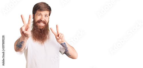 Handsome young red head man with long beard wearing casual white tshirt smiling looking to the camera showing fingers doing victory sign. number two.