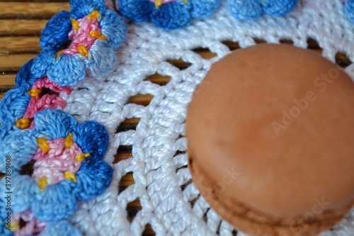Closeup of a macaron on a white crochet doily with a blue and pink flower trim on a wooden table