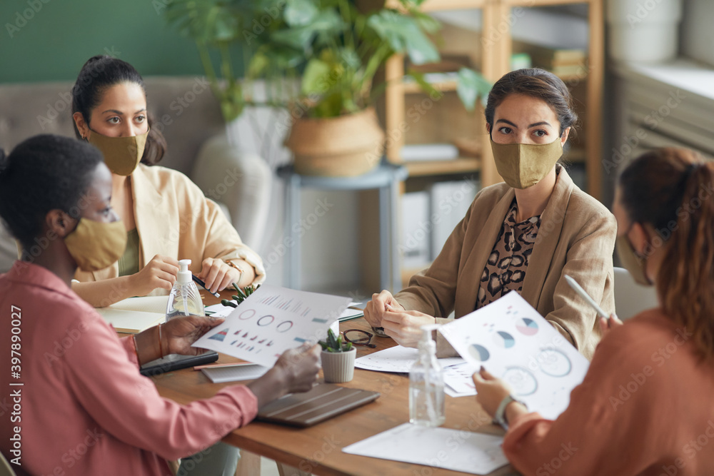 Fototapeta premium High angle view at multi-ethnic group of young businesswomen wearing masks during meeting in office, copy space
