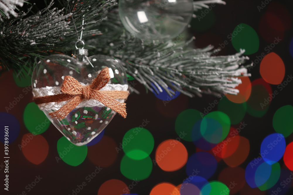 Obraz premium Heart shaped holiday bauble hanging on Christmas tree against blurred festive lights, closeup. Space for text