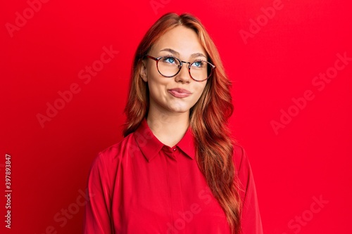 Young beautiful redhead woman wearing casual clothes and glasses over red background smiling looking to the side and staring away thinking.