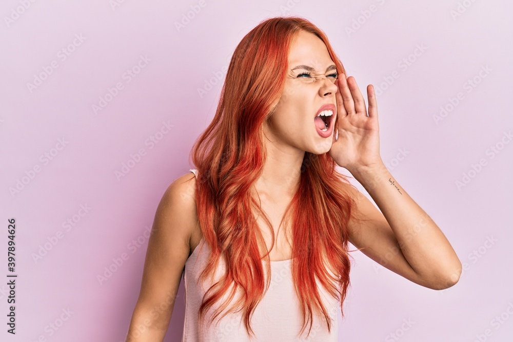 Young beautiful redhead woman wearing casual clothes over pink background shouting and screaming loud to side with hand on mouth. communication concept. Young beautiful redhead woman wearing casual clothes over pink background shouting and screaming loud to side with hand on mouth. communication concept.