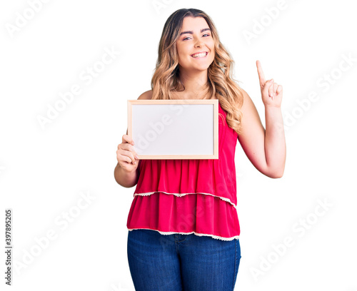 Young caucasian woman holding empty white chalkboard surprised with an idea or question pointing finger with happy face, number one