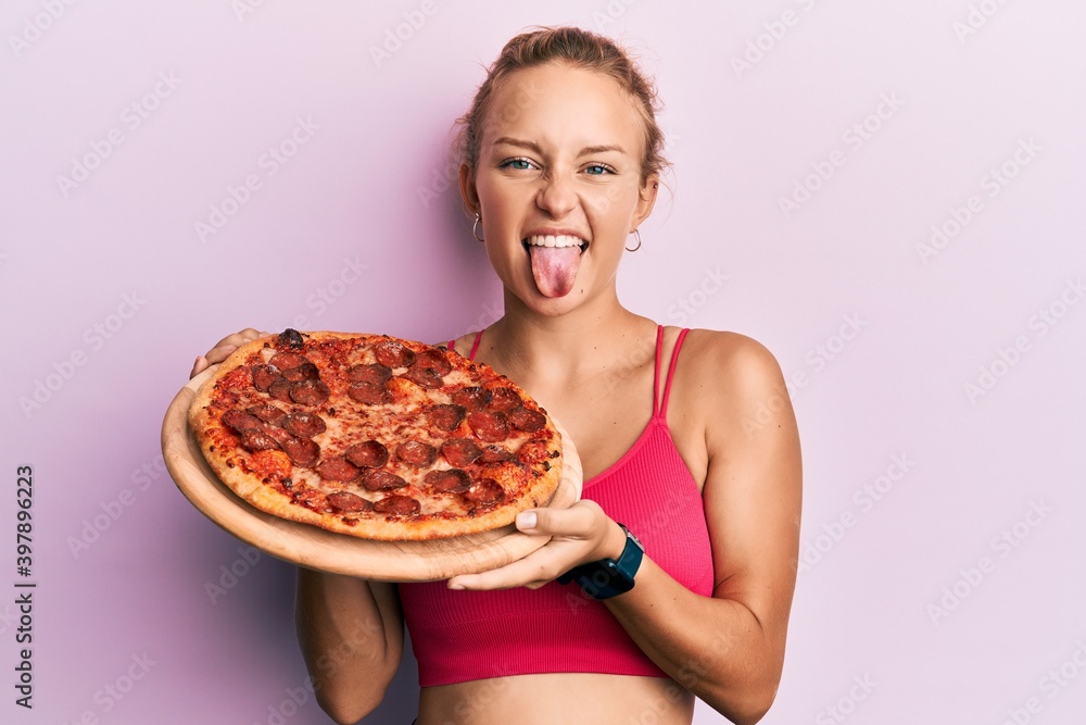 Beautiful caucasian woman holding italian pizza sticking tongue out happy with funny expression.