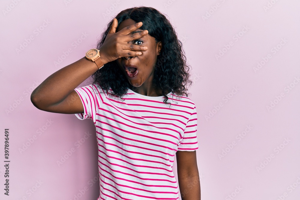Beautiful african young woman wearing casual striped t shirt peeking in shock covering face and eyes with hand, looking through fingers afraid Beautiful african young woman wearing casual striped t shirt peeking in shock covering face and eyes with hand, looking through fingers afraid