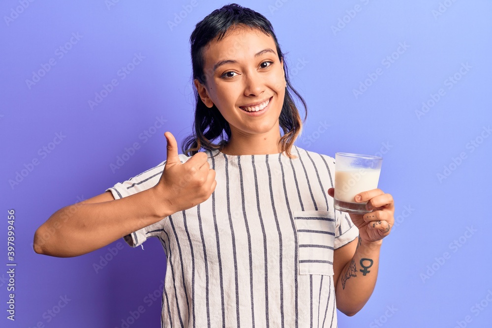 Young woman holding glass of milk smiling happy and positive, thumb up doing excellent and approval sign Young woman holding glass of milk smiling happy and positive, thumb up doing excellent and approval sign