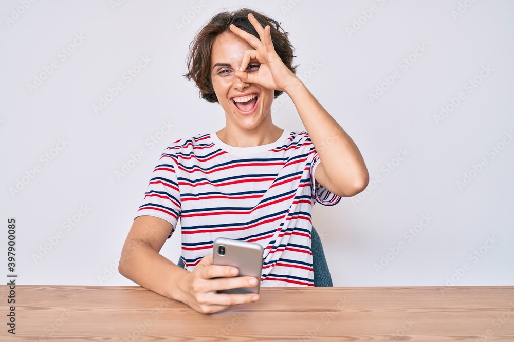 Young hispanic woman using smartphone sitting on the table smiling happy doing ok sign with hand on eye looking through fingers Young hispanic woman using smartphone sitting on the table smiling happy doing ok sign with hand on eye looking through fingers