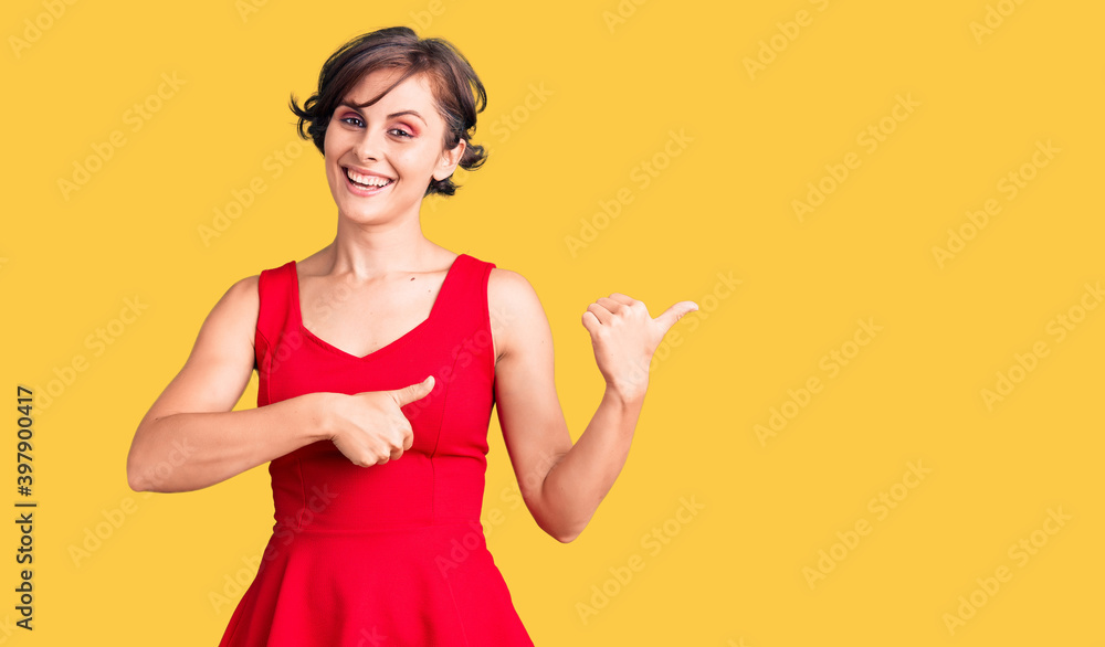 Beautiful young woman with short hair wearing casual style with sleeveless shirt pointing to the back behind with hand and thumbs up, smiling confident Beautiful young woman with short hair wearing casual style with sleeveless shirt pointing to the back behind with hand and thumbs up, smiling confident