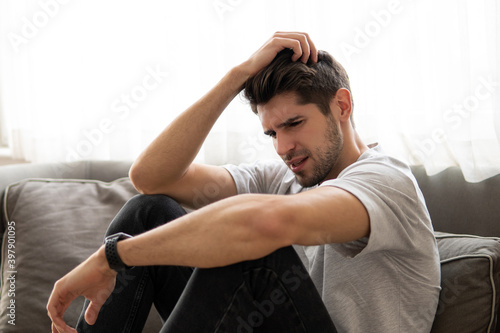 Young man with a headache is sitting on the sofa holding his head  © SlavaSelfStudio