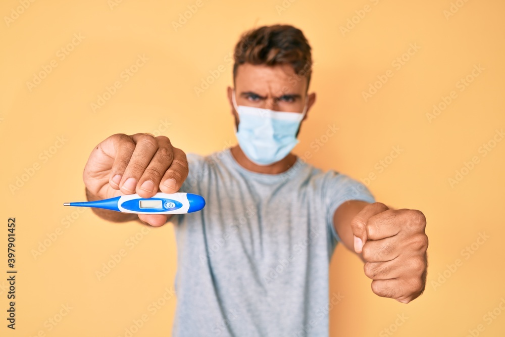 Young hispanic man wearing medical mask holding thermometer annoyed and frustrated shouting with anger, yelling crazy with anger and hand raised Young hispanic man wearing medical mask holding thermometer annoyed and frustrated shouting with anger, yelling crazy with anger and hand raised