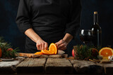Professional chef cuts fresh orange on wooden board for preparing mulled wine on rustic wooden table with festive composition background. Backstage of cooking hot drink with fragrant spices.