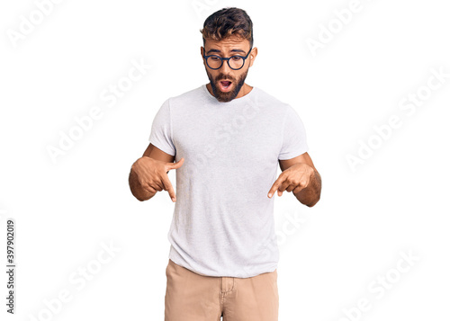 Young hispanic man wearing casual clothes and glasses pointing down with fingers showing advertisement, surprised face and open mouth