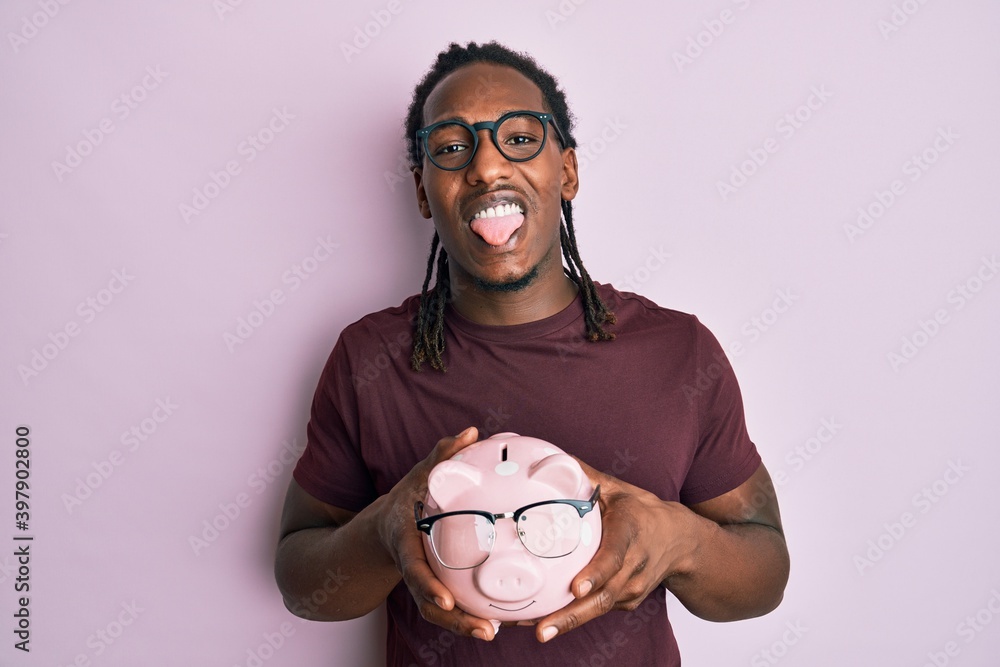 African american man with braids holding piggy bank with glasses sticking tongue out happy with funny expression. African american man with braids holding piggy bank with glasses sticking tongue out happy with funny expression.