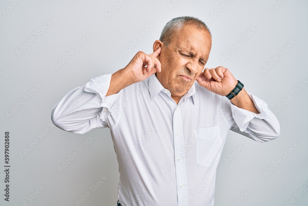 Handsome senior man wearing casual white shirt covering ears with fingers with annoyed expression for the noise of loud music. deaf concept. Handsome senior man wearing casual white shirt covering ears with fingers with annoyed expression for the noise of loud music. deaf concept.