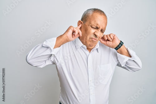 Handsome senior man wearing casual white shirt covering ears with fingers with annoyed expression for the noise of loud music. deaf concept. © Krakenimages.com
