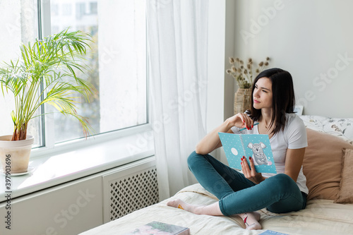 Brunette girl sitting on the bed in a bright room reading a book.