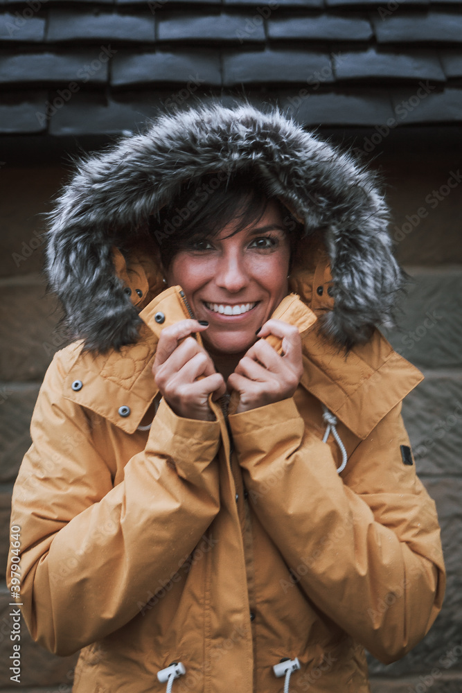 Winter portrait of a beautiful woman smiling with her fur hood on while holding the neckline of her yellow jacket Winter portrait of a beautiful woman smiling with her fur hood on while holding the neckline of her yellow jacket