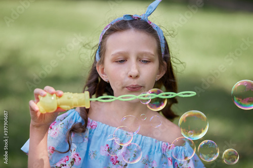 A beautiful girl sends soap bubbles. Looks at the camera. Summer Park. The dress and bow on the head are blue.