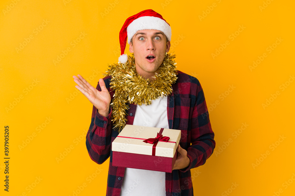Young caucasian man with christmas hat holding a present isolated on yellow background Young caucasian man with christmas hat holding a present isolated on yellow background