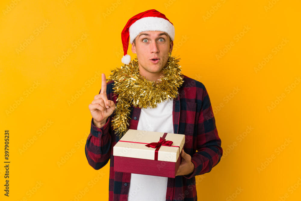 Young caucasian man with christmas hat holding a present isolated on yellow background Young caucasian man with christmas hat holding a present isolated on yellow background