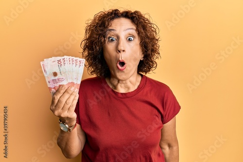 Beautiful middle age mature woman holding 10 colombian pesos banknotes scared and amazed with open mouth for surprise, disbelief face