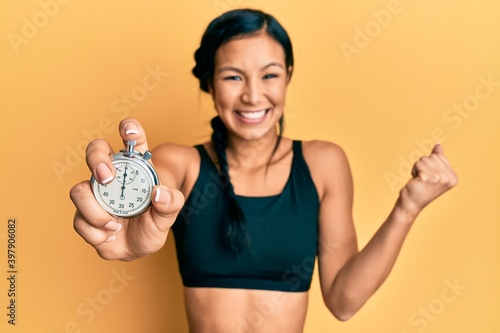 Beautiful hispanic woman wearing sportswear holding stopwatch screaming proud  celebrating victory and success very excited with raised arm