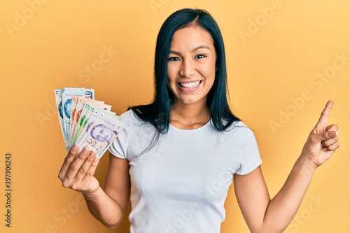 Beautiful hispanic woman holding singapore dollars banknotes smiling happy pointing with hand and finger to the side