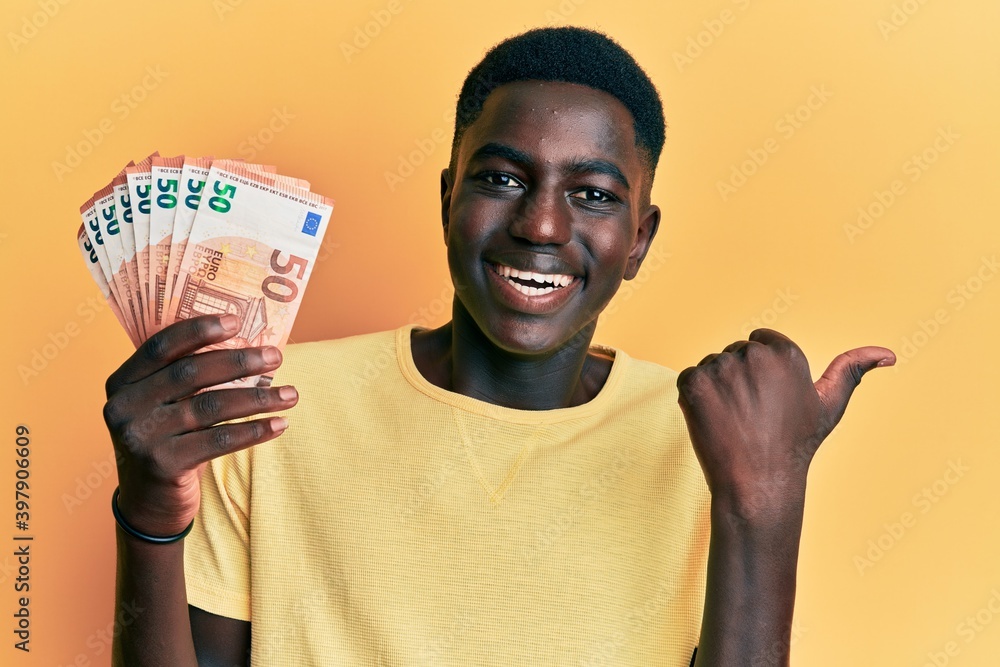 Young african american man holding 50 euro banknotes pointing thumb up to the side smiling happy with open mouth Young african american man holding 50 euro banknotes pointing thumb up to the side smiling happy with open mouth