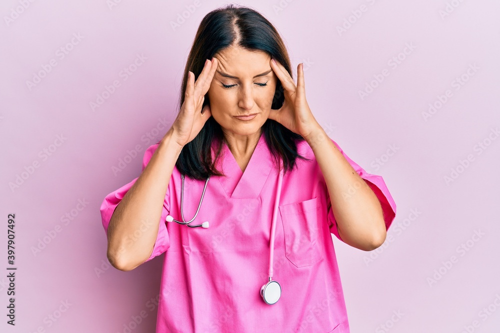 Middle age brunette woman wearing doctor uniform and stethoscope with hand on head for pain in head because stress. suffering migraine. Middle age brunette woman wearing doctor uniform and stethoscope with hand on head for pain in head because stress. suffering migraine.
