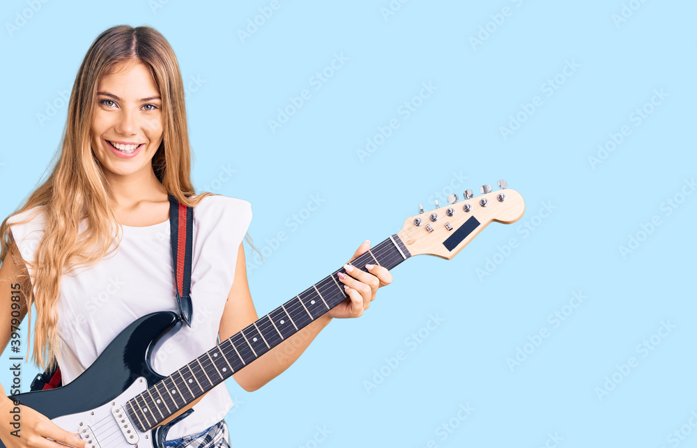 Fototapeta premium Beautiful caucasian woman with blonde hair playing electric guitar looking positive and happy standing and smiling with a confident smile showing teeth