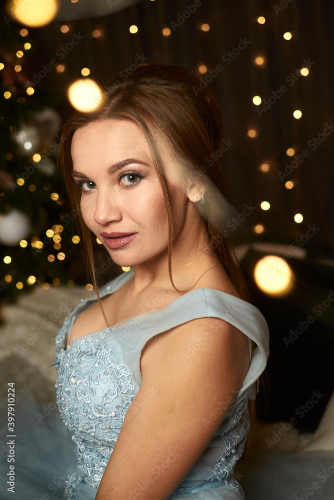 Fototapeta premium Beautiful woman in a blue evening dress on the background of a Christmas tree and garlands. Holiday concept, new year, christmas, sale and discounts.