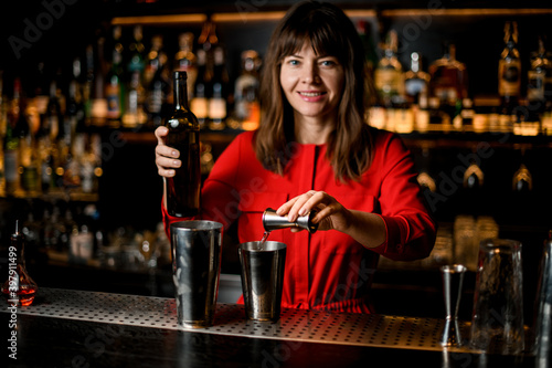 smiling woman bartender pours liquid from jigger into steel shaker cup standing on bar