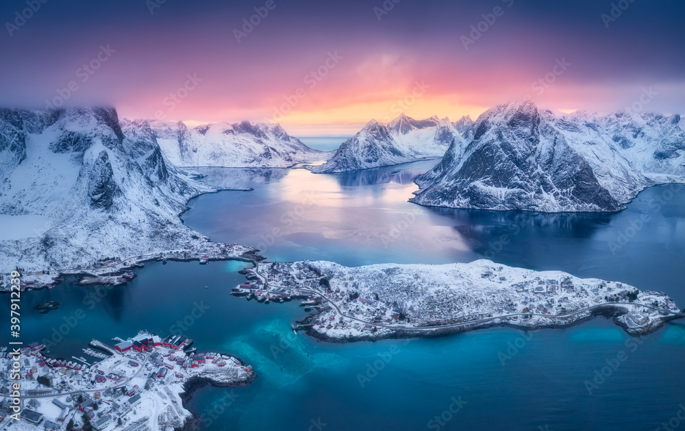 Aerial view of Reine at sunset in winter. Top view of Lofoten islands, Norway. Landscape with blue sea, snowy mountains, high rocks, village with buildings, rorbu, colorful sky, reflection in water