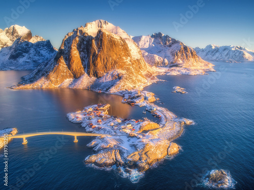 Aerial view of Hamnoy at sunrise in winter. Lofoten islands, Norway. Panoramic colorful landscape with blue sea, snowy mountains, islands, rocks, village, rorbu, road, bridge, sky. Top view from drone